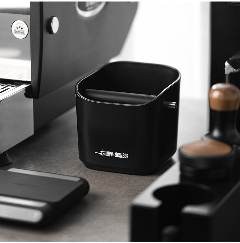 Knock box MHW-3BOMBER Square en noir (Black) avec bord biseauté à côté d’une machine espresso
