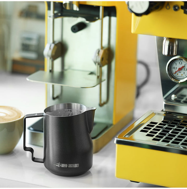 Pichet à lait MHW-3BOMBER Turbo en noir mat (Matte Black) avec poignée galbée à côté d’une machine espresso