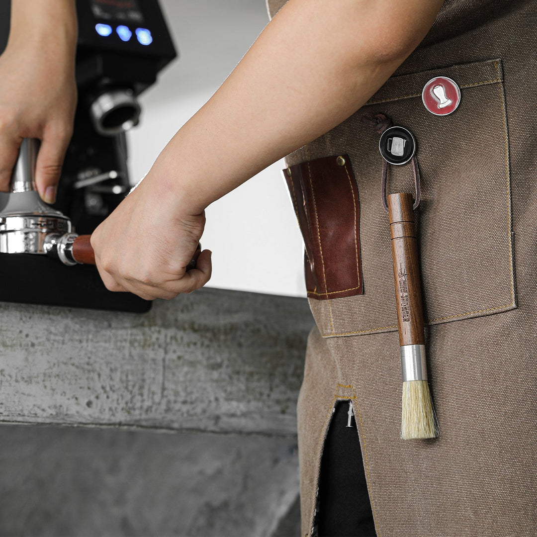 Pinceau barista MHW-3BOMBER Wings Cylindrical Brush en noyer (Walnut) à portée de main sur un tablier de barista