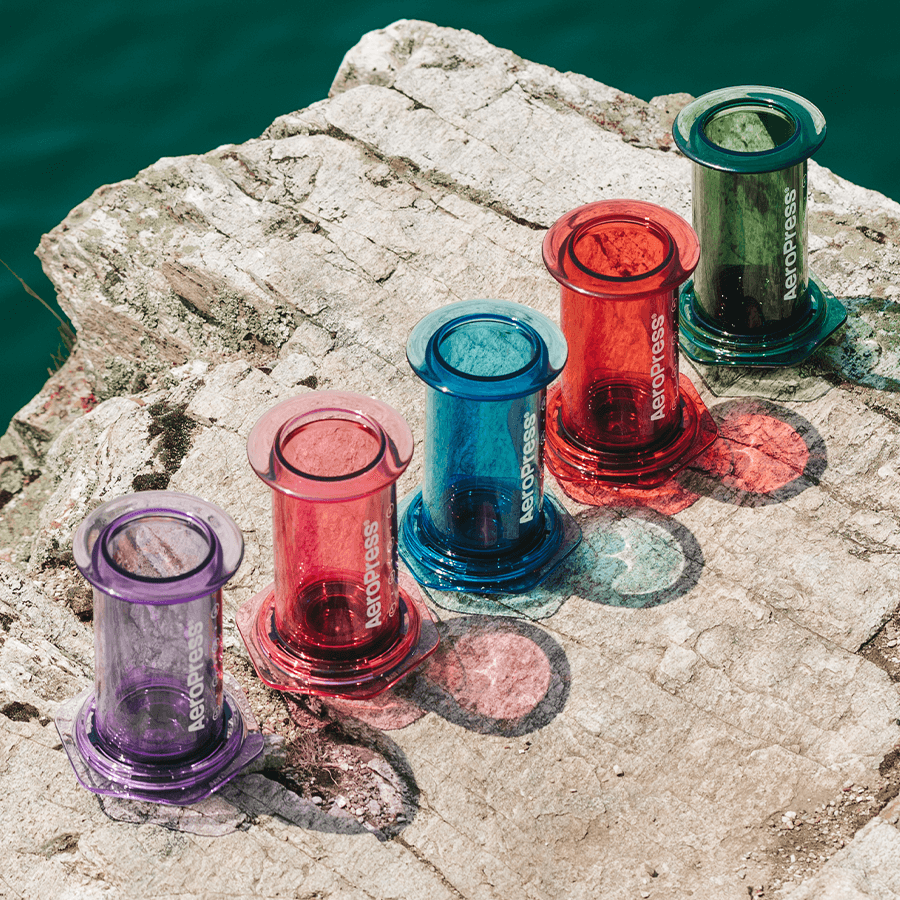AeroPress Clear in Violett, Rosa, Blau, Rot und Grün auf einem Felsen am Wasser – Coffee Coaching Club 24