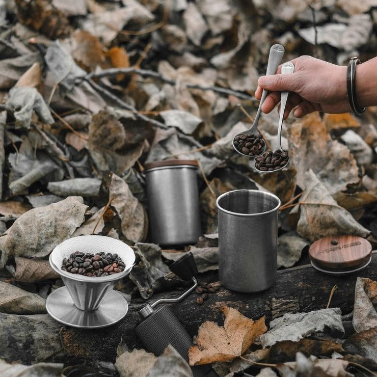 Boîte en inox MHW-3BOMBER en gris argenté (Silver Spot) avec couvercle en bois à côté d’un dripper et de grains de café