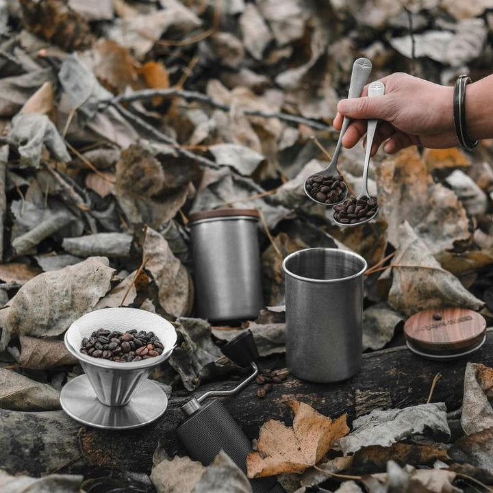 Boîte en inox MHW-3BOMBER en gris argenté (Silver Spot) avec couvercle en bois à côté d’un dripper et de grains de café